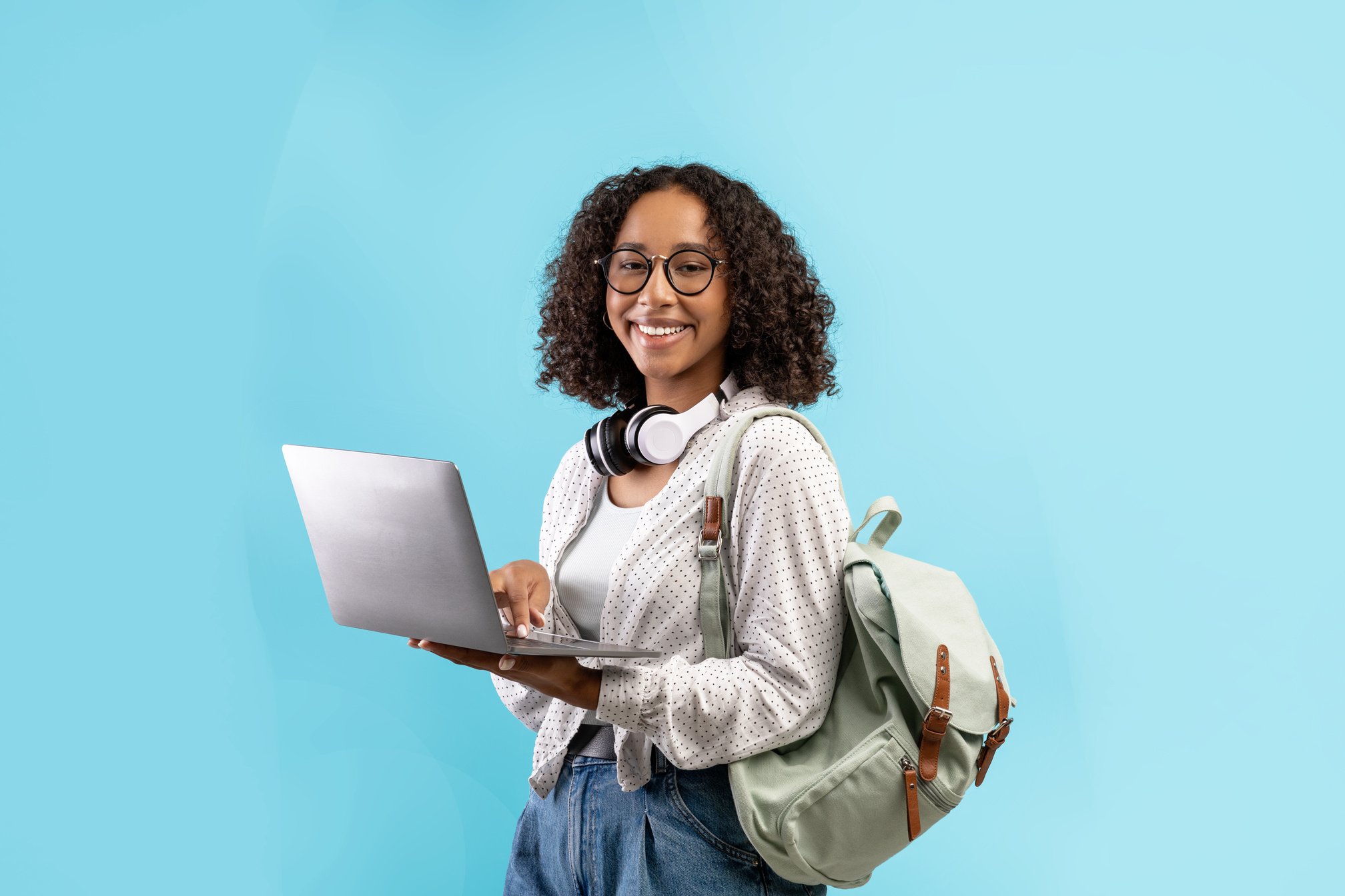 Cheerful young black woman with backpack and headphones holding laptop pc over blue studio background